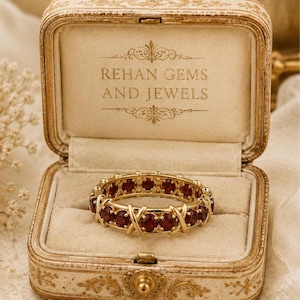 Könnte beinhalten: Ein goldener Eternity-Ring mit runden, tiefroten Granat-Edelsteinen, die in einem sich wiederholenden X-förmigen Metallmuster gefasst sind. Der Ring liegt in einer cremefarbenen, rechteckigen Schmuckschatulle mit dem Aufdruck REHAN GEMS AND JEWELS.