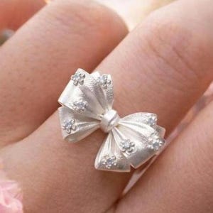 May include: A silver-toned ring featuring a prominent bow-shaped centerpiece. The bow has a textured, matte finish and is adorned with several small, clear, sparkling stones arranged in a floral pattern across the surface of the bow loops.