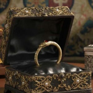 May include: A gold-colored ring in the shape of a snake with a red gemstone eye, displayed in an open black jewelry box. The box is decorated with gold filigree and pearl accents, creating a luxurious aesthetic.