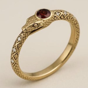Könnte beinhalten: Ein goldfarbener Ring in Form einer Schlange. Der Schlangenkopf hat einen runden, roten Edelstein und das Band ist mit kleinen Diamanten verziert. Der Ring hat ein strukturiertes, schuppenartiges Muster.