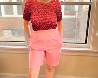 Puede incluir: Un conjunto de dos piezas con una parte superior roja texturizada de manga corta y pantalones cortos rosas de cintura alta. La parte superior tiene una textura única y burbujeante, mientras que los pantalones cortos tienen un ajuste a medida con bolsillos. El atuendo es usado.