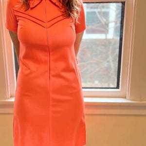Puede incluir: Un vestido naranja brillante con mangas cortas y cuello. El vestido tiene una cremallera de largo completo y costuras decorativas en el pecho. El vestido, a la altura de la rodilla, tiene una silueta ajustada.