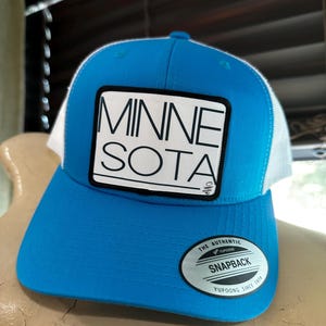 May include: A blue and white trucker hat with a black and white patch that says "MINNESOTA" on it. The hat has a white mesh back and a black "Yupoong" label on the side.