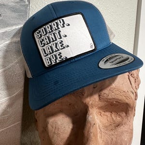 May include: A blue and gray trucker hat with a white patch that says "Sorry, Can't, Lake, Bye."