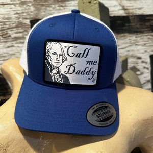 May include: A blue and white trucker hat with a patch that says "Call me Daddy" and a picture of George Washington. The hat has a white mesh back and a black "The Authentic Snapback Yupoong Since 1974" label.