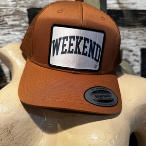 May include: A brown and white trucker hat with a white patch that says "WEEKEND" in black letters. The hat has a mesh back and a black snap closure. The hat also has a silver label that says "The Authentic Snapback YUPOONG Since 1974."