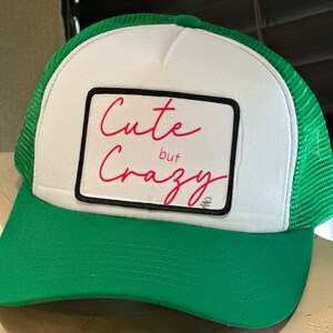 May include: A white and green trucker hat with a black and white patch that says "Cute but Crazy" in red cursive writing.