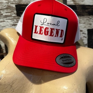 May include: Red and white mesh trucker hat with a white patch that says "Local Legend" in black script. The hat has a black and silver "The Authentic Snapback" label.