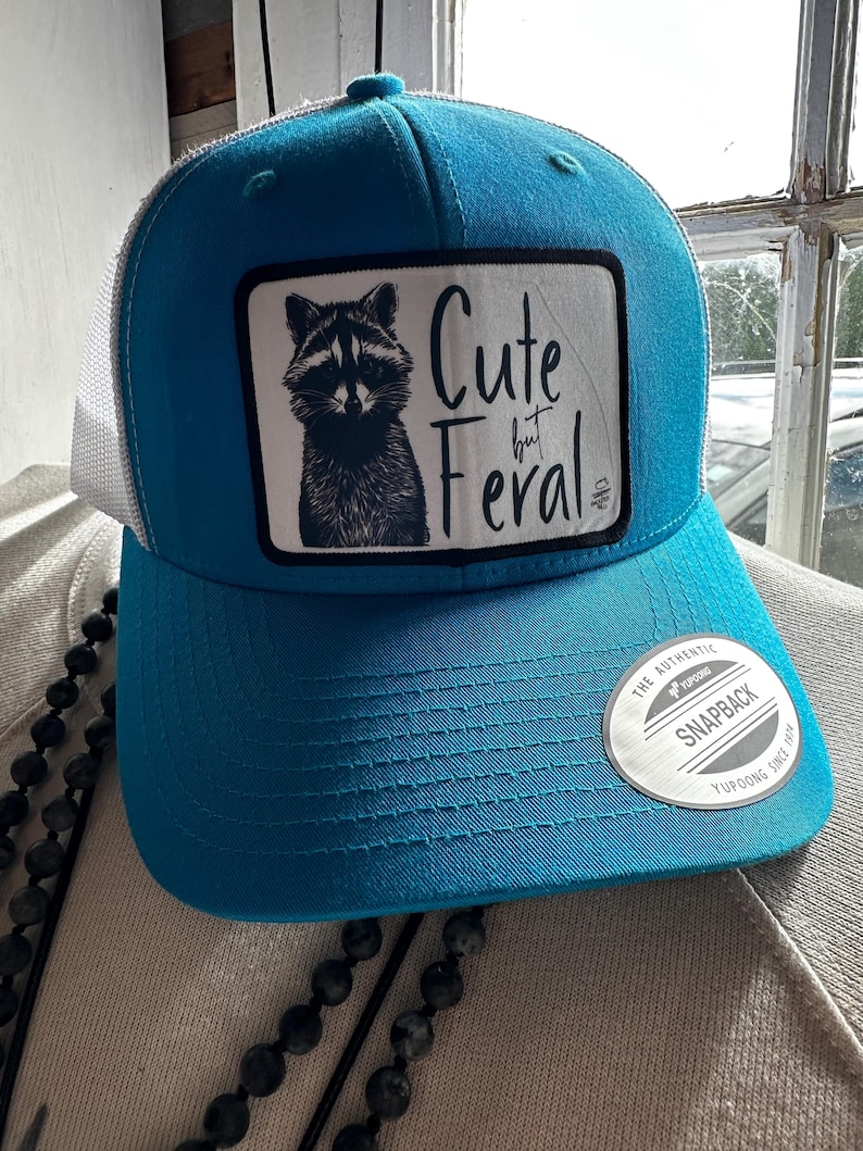 以下が含まれることがあります： ターコイズブルーと白のトラッカーハット。黒と白のラクーンドローイングと「Cute but Feral」の文字がデザインされています。ハットは白のメッシュバックと黒の調節可能なストラップが付いています。