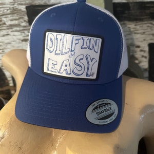 May include: A blue and white trucker hat with a black patch that says "Dilfin Ain't Easy". The hat has a white mesh back and a black "The Authentic Yupoong Snapback Since 1974" label.