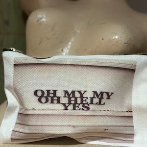 Könnte beinhalten: Eine weiße Canvas-Reißverschlusstasche mit einem braunen und weißen Streifenmuster. Die Tasche hat den Text "OH MY MY OH HELL YES" in Schwarz aufgedruckt.