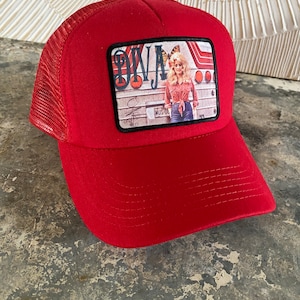 May include: Red trucker hat with a black patch featuring a photo of a woman with blonde hair and a butterfly. The patch also has the text "ONJA" and "MUS-OL".