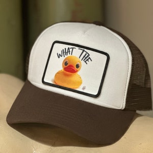 May include: A white and brown trucker hat with a yellow rubber ducky and the text "WHAT THE" on a black patch.