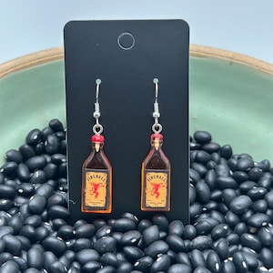 Op de afbeelding: Oorbellen met Fireball-flesjes en zilveren haken. Elke oorbel heeft een miniatuurreplica van een Fireball-kaneelwhiskeyflesje met een rode dop en een etiket met een rode draak. Gepresenteerd tegen een zwarte achtergrond.