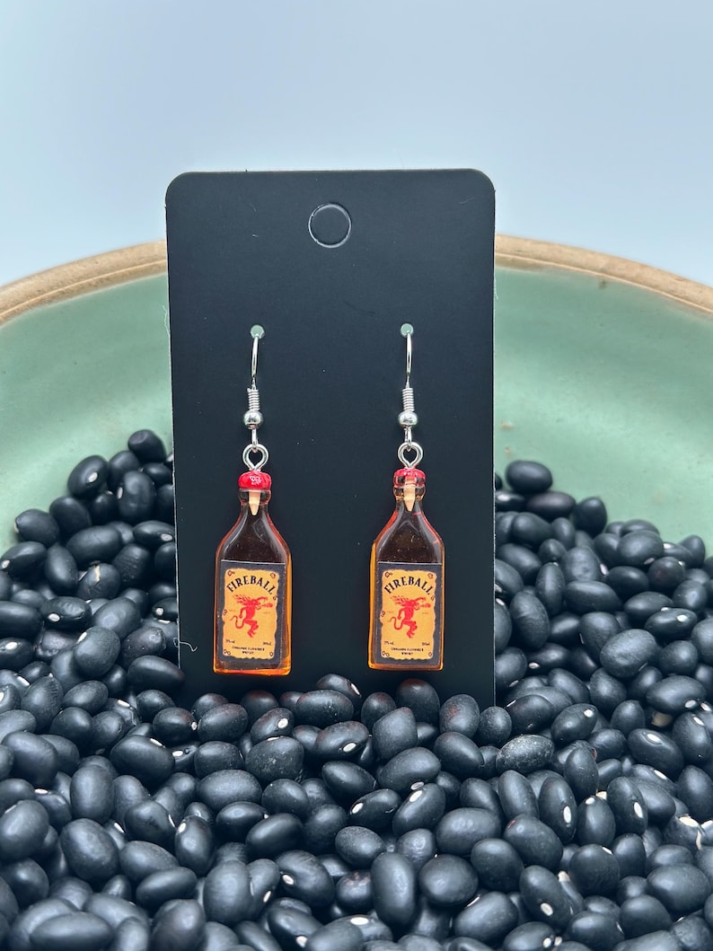 May include: Fireball bottle earrings with silver hooks. Each earring features a miniature replica of a Fireball cinnamon whiskey bottle with a red cap and a label with a red dragon. The earrings are displayed against a black background.
