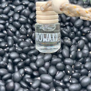 Pode incluir: Um frasco difusor de ar de vidro transparente com o nome "HOWARD" impresso, coberto com uma tampa de madeira e uma corda. A garrafa é cercada por uma cama de feijão preto.
