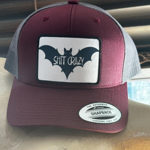 May include: A burgundy and gray trucker hat with a black and white patch that says "Shit Crazy" and a black bat silhouette. The hat has a black and white "The Authentic Yupoong Snapback Yupoong Since 1974" label.