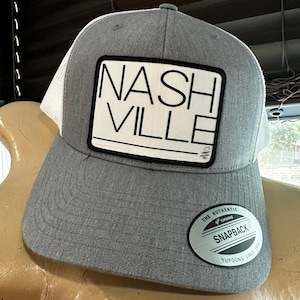 May include: Gray and white mesh baseball cap with a black patch that says "NASHVILLE". The cap has a black and white sticker that says "THE AUTHENTIC YUPOONG SNAPBACK YUPOONG SINCE 1974".