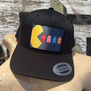 May include: Black trucker hat with a yellow Pac-Man and four ghosts in blue, pink, red, and orange on the front. The hat has a black mesh back and a white "The Authentic Yupoong Snapback Yupoong Since 1974" label.