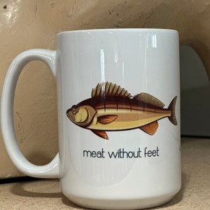 May include: White ceramic coffee mug with a brown and white fish illustration and the text "meat without feet".