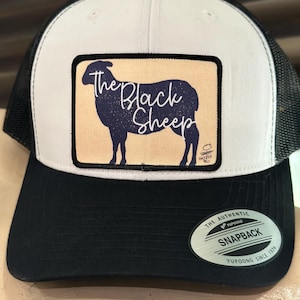May include: A white and black trucker hat with a black sheep silhouette and the text "The Black Sheep" on a tan patch. The hat has a black mesh back and a black YUPOONG SNAPBACK label.