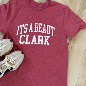 May include: A red heather t-shirt with white text that reads "IT'S A BEAUT CLARK".
