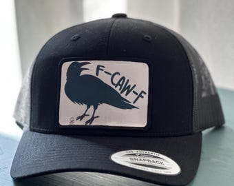 May include: Black trucker hat with a mesh back and a curved brim. The front features a rectangular patch with a crow silhouette and the text "F-CAW-F" in white. The hat has a snapback closure.