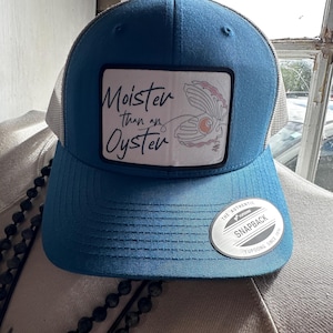 May include: A blue and white trucker hat with a white patch that says "Moister than an Oyster" and a drawing of an oyster with a pearl inside. The hat has a white mesh back and a black "The Authentic Snapback Yupoong Since 1974" label.