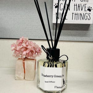 May include: A clear, hexagonal glass reed diffuser labeled "Strawberry Green Tea" with black reeds. The label includes a cat silhouette. A small sign reads "I WORK HARD SO MY CAT CAN HAVE NICE THINGS." Pink flowers and two white wrapped objects are also present.