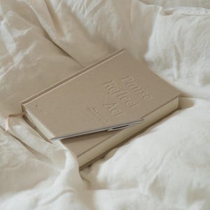 Puede incluir: Un diario beige con el texto en relieve "Pause Reflect Act" y un bol&iacute;grafo plateado encima. El diario est&aacute; sobre una cama con s&aacute;banas blancas. El diario tiene una cinta de marcador.
