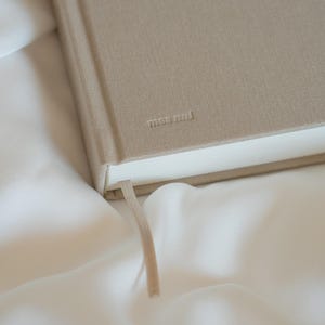 Puede incluir: Un diario beige con el texto en relieve "THE JOURNEY OF A GRATEFUL HEART" y la marca "mae&mini". El diario tiene una cinta de marcador beige a juego y est&aacute; sobre una superficie blanca y sedosa.