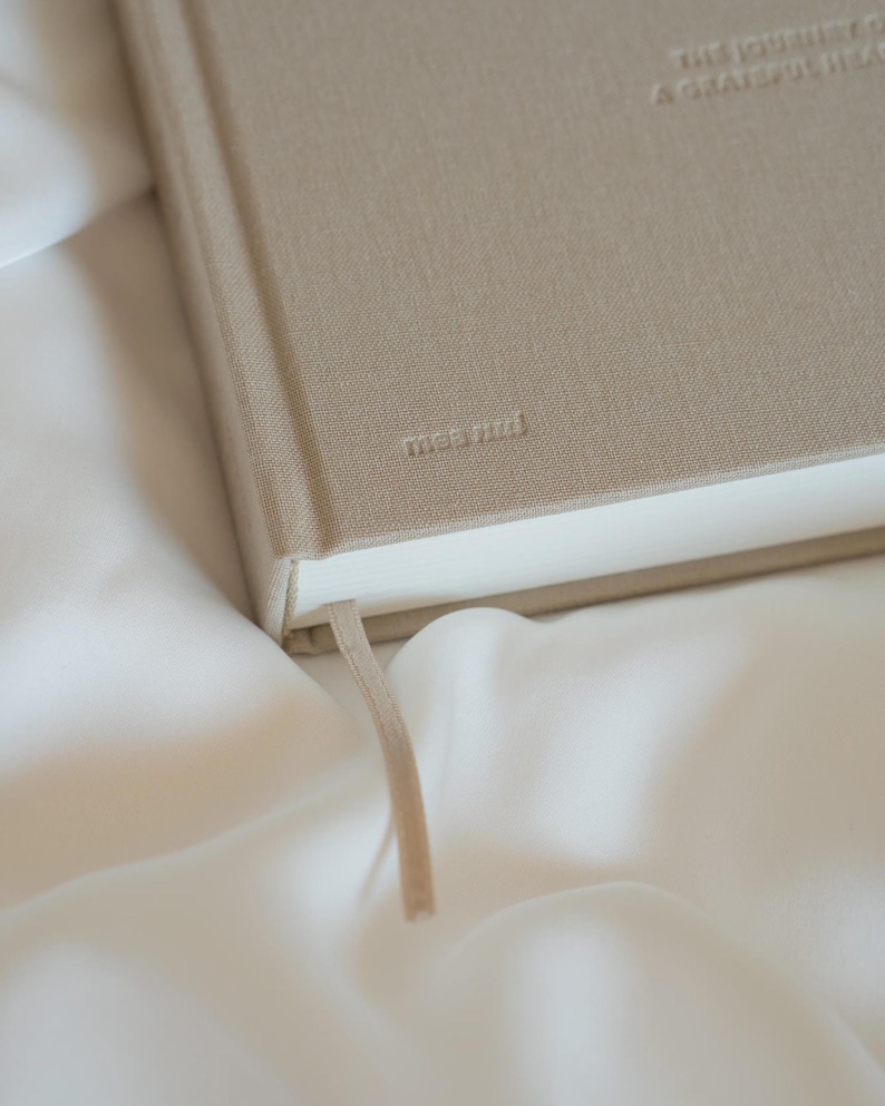 Puede incluir: Un diario beige con el texto en relieve "THE JOURNEY OF A GRATEFUL HEART" y la marca "mae&mini". El diario tiene una cinta de marcador beige a juego y est&aacute; sobre una superficie blanca y sedosa.
