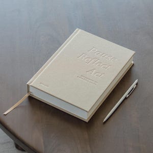 Puede incluir: Un diario beige con las palabras en relieve "Pause Reflect Act" y un marcap&aacute;ginas de cinta a juego. Un bol&iacute;grafo plateado descansa junto al diario sobre una superficie de madera. El diario tambi&eacute;n tiene el texto "The Journal of a Grateful Heart".