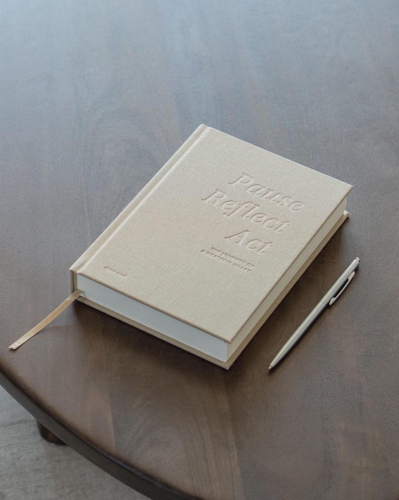 Puede incluir: Un diario beige con las palabras en relieve "Pause Reflect Act" y un marcap&aacute;ginas de cinta a juego. Un bol&iacute;grafo plateado descansa junto al diario sobre una superficie de madera. El diario tambi&eacute;n tiene el texto "The Journal of a Grateful Heart".