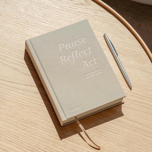 Könnte beinhalten: Ein hellbeiges Notizbuch mit festem Einband und dem geprägten Text "Pause Reflect Act" sowie "The Journey of a Grateful Heart" auf dem Vorderdeckel, ergänzt durch einen schlanken silbernen Metallstift und ein beiges Lesebändchen.
