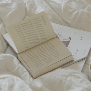 Puede incluir: Un libro abierto y una revista con la palabra "ARK" sobre una cama blanca. Las p&aacute;ginas del libro est&aacute;n llenas de texto. Un bol&iacute;grafo descansa sobre la revista. La revista tiene las palabras "JOUR" y "SPACES".