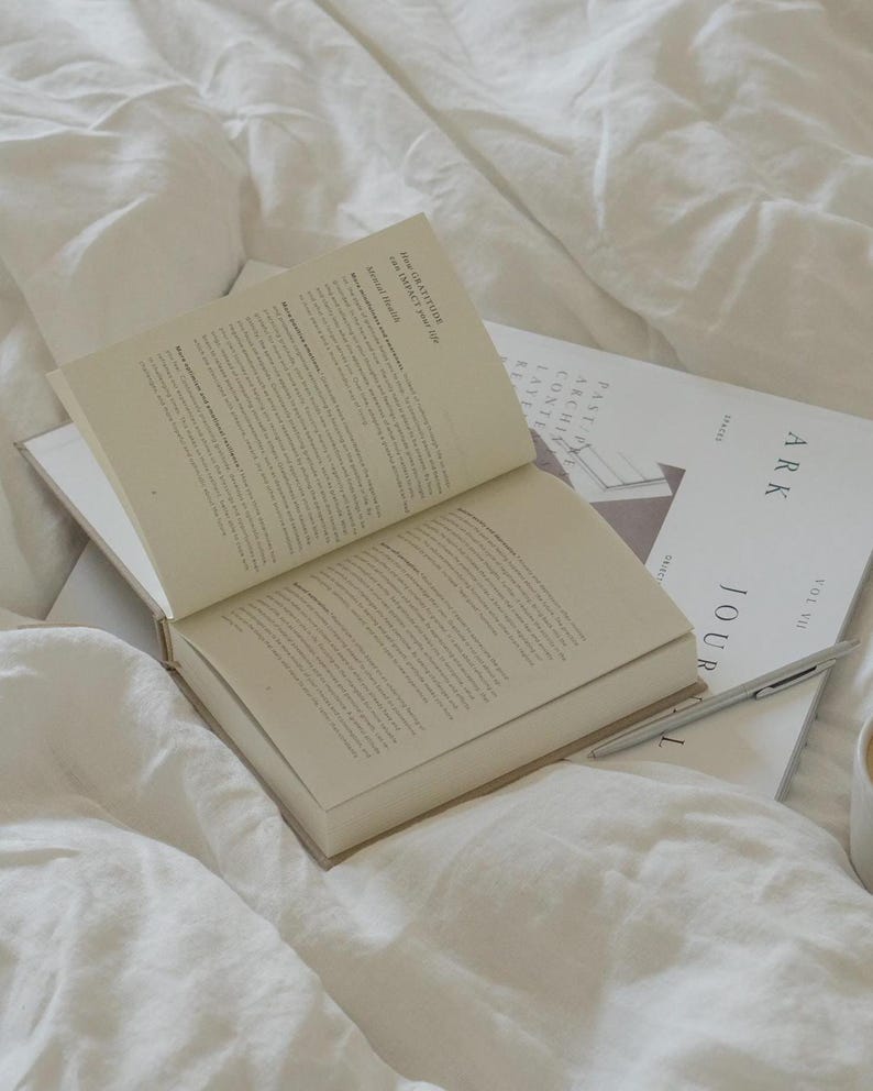 Puede incluir: Un libro abierto y una revista con la palabra "ARK" sobre una cama blanca. Las p&aacute;ginas del libro est&aacute;n llenas de texto. Un bol&iacute;grafo descansa sobre la revista. La revista tiene las palabras "JOUR" y "SPACES".