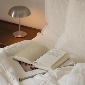 Puede incluir: Una peque&ntilde;a l&aacute;mpara de escritorio plateada con una pantalla redondeada est&aacute; sobre una superficie de madera junto a una cama. Un libro abierto y una revista descansan sobre la ropa de cama blanca. La escena est&aacute; ba&ntilde;ada en luz suave y c&aacute;lida.