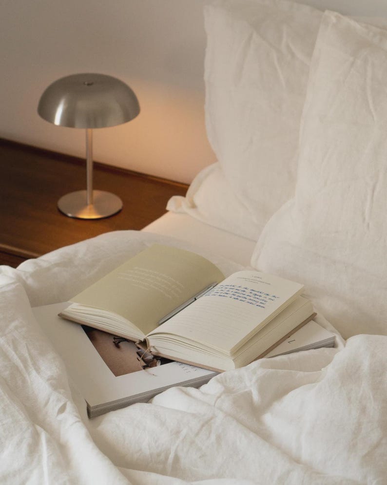 Puede incluir: Una peque&ntilde;a l&aacute;mpara de escritorio plateada con una pantalla redondeada est&aacute; sobre una superficie de madera junto a una cama. Un libro abierto y una revista descansan sobre la ropa de cama blanca. La escena est&aacute; ba&ntilde;ada en luz suave y c&aacute;lida.
