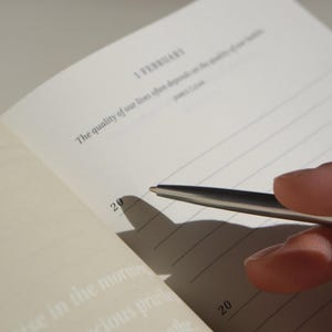 Puede incluir: Cuaderno abierto con p&aacute;ginas rayadas, con el texto "1 FEBRERO" y una cita. Un bol&iacute;grafo plateado se cierne sobre la p&aacute;gina, proyectando una sombra. El n&uacute;mero "20" es visible. Las p&aacute;ginas del cuaderno son de color blanquecino.