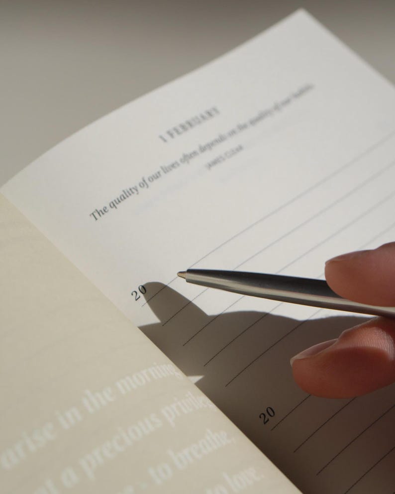 Puede incluir: Cuaderno abierto con p&aacute;ginas rayadas, con el texto "1 FEBRERO" y una cita. Un bol&iacute;grafo plateado se cierne sobre la p&aacute;gina, proyectando una sombra. El n&uacute;mero "20" es visible. Las p&aacute;ginas del cuaderno son de color blanquecino.