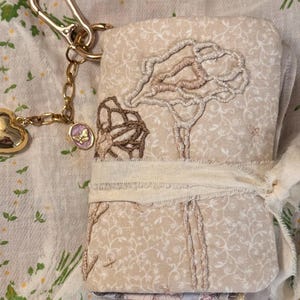 Può includere: Un oggetto rivestito in tessuto beige con ricami di alberi e motivi floreali, legato con un nastro color crema. Un portachiavi color oro con un ciondolo a forma di cuore e un piccolo pendente con un disegno a farfalla è attaccato.