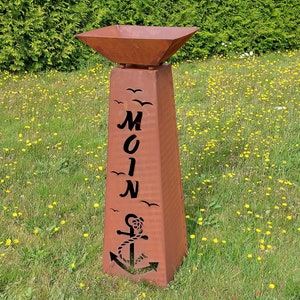 Könnte beinhalten: Ein rostiger Metall-Feuertischständer mit quadratischer Oberseite und einem ausgeschnittenen Design eines Ankers, Möwen und dem Wort "MOIN".