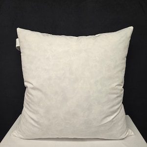 Op de afbeelding: Een wit, vierkant kussen met een zachte, gladde textuur. Het kussen ligt op een wit oppervlak.