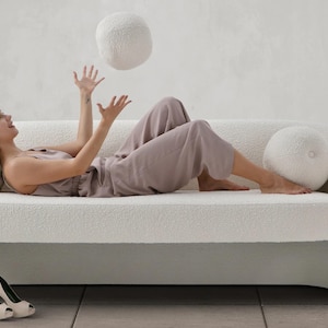 May include: A white, fluffy sofa with a woman lying on it, tossing a white ball in the air. The sofa has rounded edges and a plush, textured fabric.