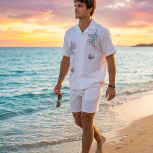 Könnte beinhalten: Weißes Leinenhemd und Shorts-Set mit gestickten Blumen- und Schmetterlingsdetails. Das kurzärmelige Hemd hat einen Hemdkragen. Das Outfit wird am Strand bei Sonnenuntergang getragen. Die Person hält eine Sonnenbrille.