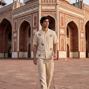 Puede incluir: Conjunto de camisa y pantal&oacute;n de lino beige con bordados florales coloridos. La camisa de manga corta tiene cuello y cierre de botones. El pantal&oacute;n tiene cintura con cord&oacute;n. El conjunto se lleva con sandalias blancas. Un gran edificio en el fondo.