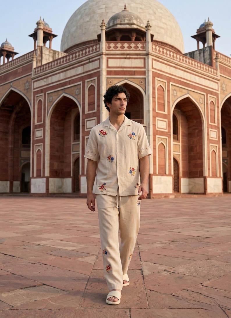 Puede incluir: Conjunto de camisa y pantal&oacute;n de lino beige con bordados florales coloridos. La camisa de manga corta tiene cuello y cierre de botones. El pantal&oacute;n tiene cintura con cord&oacute;n. El conjunto se lleva con sandalias blancas. Un gran edificio en el fondo.