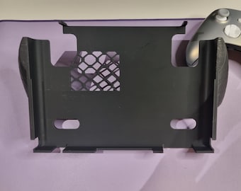 May include: A black plastic stand with a cutout for a video game console. The stand has a mesh design on the back and a small opening on the front.