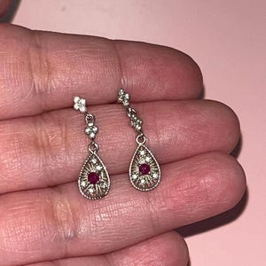 May include: A pair of silver-toned drop earrings. Each earring features a small flower at the top, a series of small clear stones, and a teardrop-shaped pendant with a central red stone and surrounding clear stones. The earrings are displayed against a pink background.